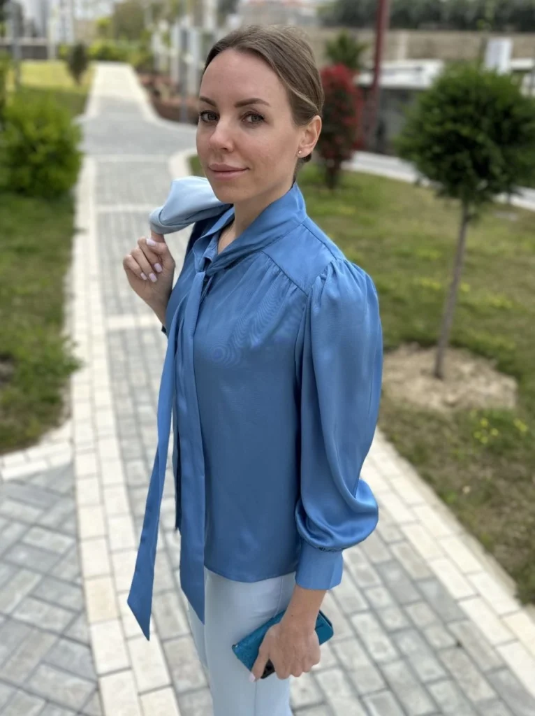 An elegant, close-up outdoor portrait of a woman wearing a light-blue satin tie-neck blouse, standing in a modern garden area with a paved path, holding a clutch and looking directly at the camera.