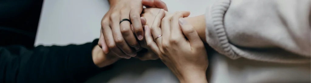 A close-up photograph of multiple hands from different people interlocked and held together on a neutral, soft surface, symbolizing unity, support, and collective strength.