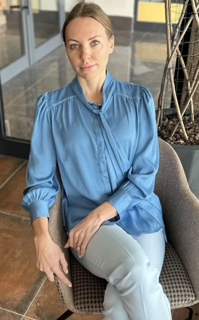 A professional, elegant close-up portrait of a woman with blonde hair, wearing a stylish, blue satin blouse with puff sleeves and a tie neck, seated comfortably in a brown velvet chair in an indoor setting.