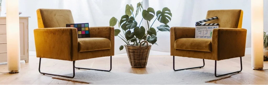 An interior photograph of a therapy or interview room with two identical plush, golden-mustard armchairs with black metal frames, facing each other. Between them is a potted Monstera plant, with soft white drapes in the background and a clapperboard on one chair.