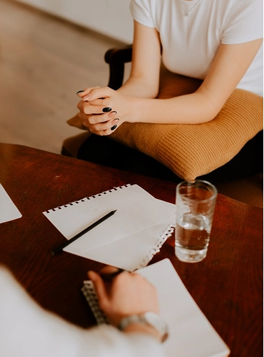 An overhead view focusing on a patient's interlocked hands resting on a pillow during a therapy session, with a therapist's hand and writing materials on a wooden table in the foreground.