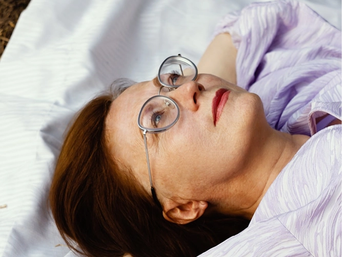 A close-up portrait of a thoughtful older woman with ginger hair wearing round, silver-rimmed glasses, looking upward and to the side, wearing a light-purple textured blouse, resting on a white sheet.