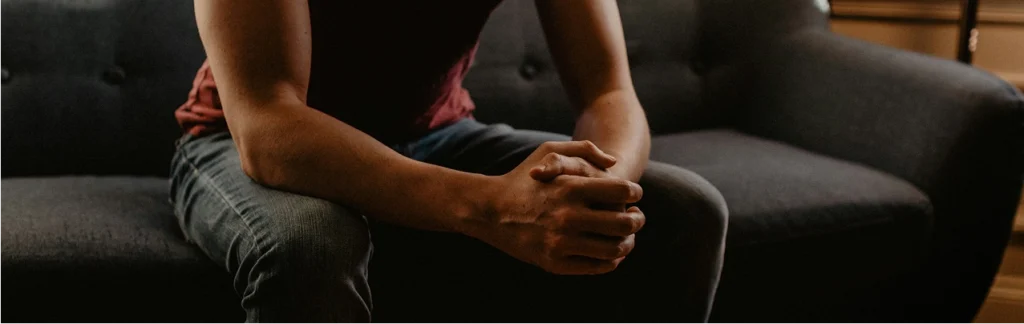 A person sits on a dark sofa, leaning forward with hands clasped, wearing a red shirt and jeans. The image conveys a sense of contemplation or tension.